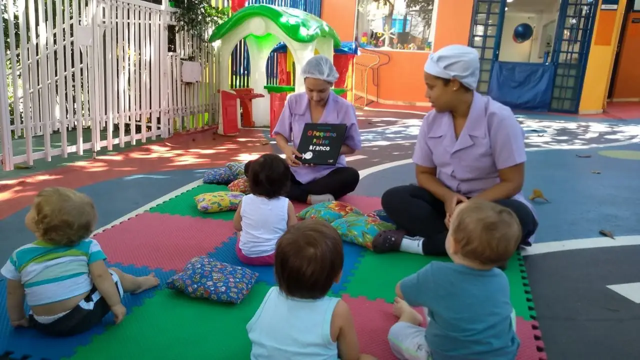 Bebês sentados em emborrachados, em uma área externa, à sombra de árvores, observando duas colaboradoras do Berçário da Escola Mundo Feliz lerem uma história.